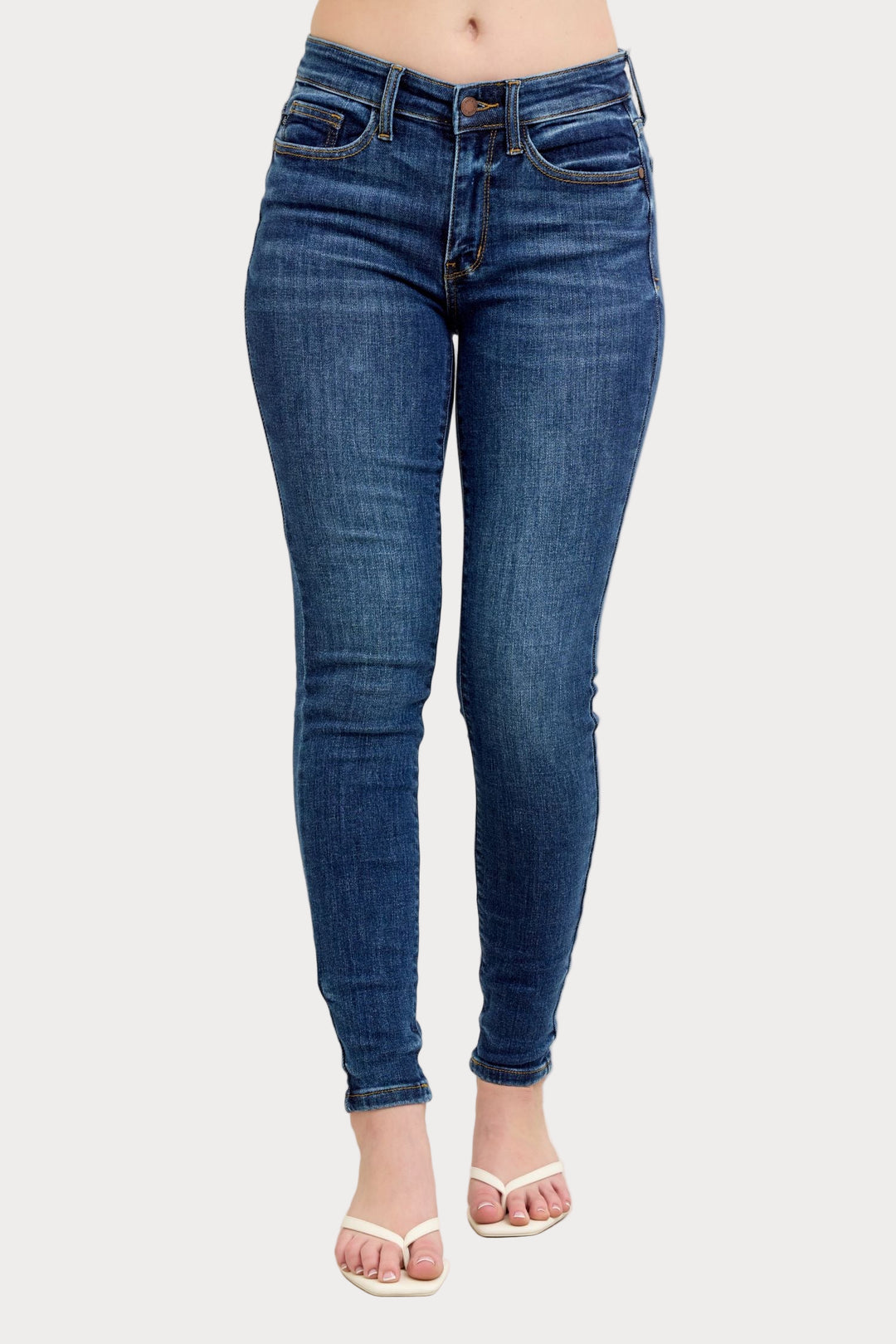 Front view of a model wearing the Anna Mid Rise Skinny Jeans, a non-distressed dark wash denim style from Judy Blue. She is standing against a light grey background and wearing white flip-flop sandals.
