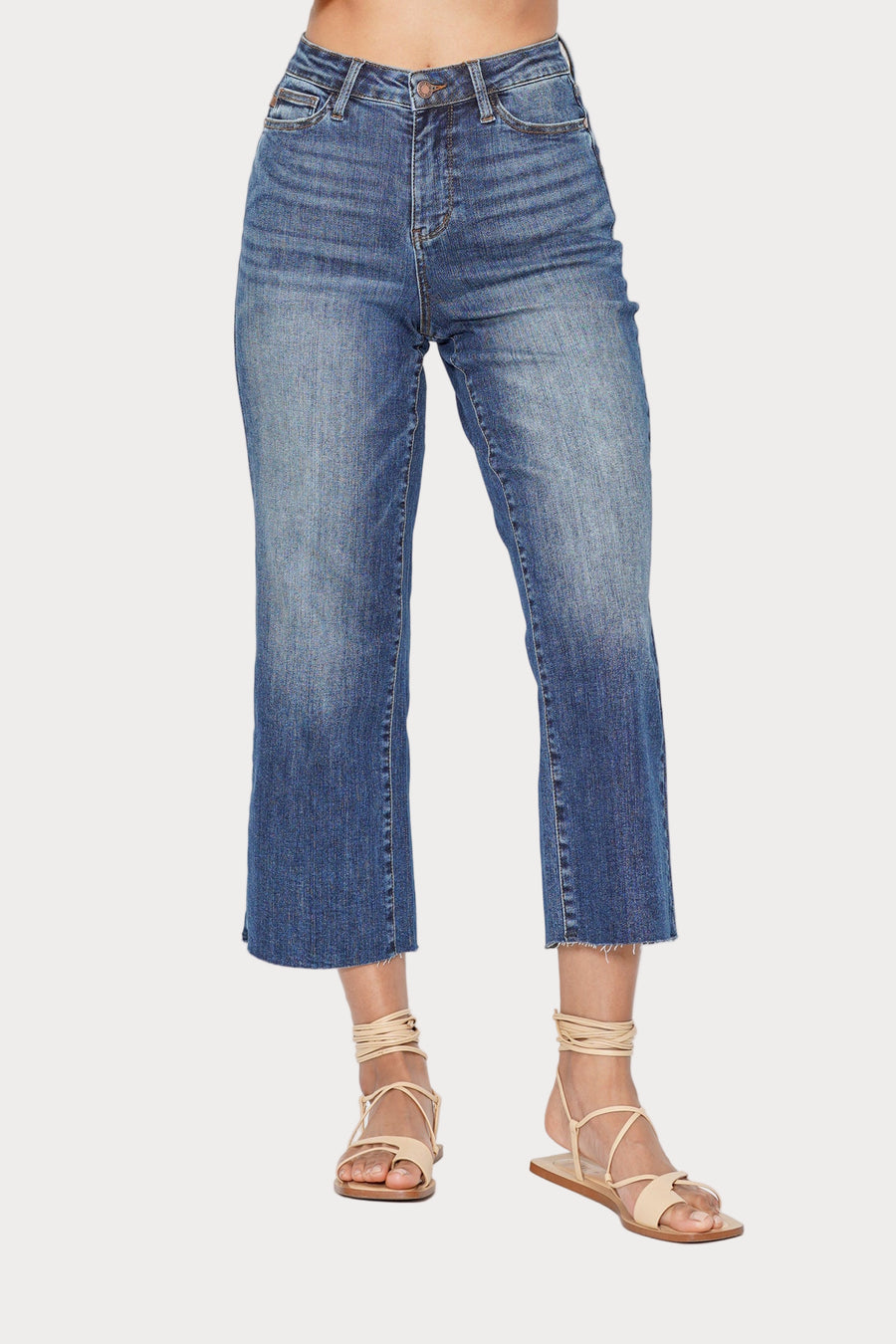 Front view of a model wearing the Dolly high-rise cropped wide-leg jeans in a medium wash with a raw hem. She is standing against a light grey background and wearing beige strappy sandals.