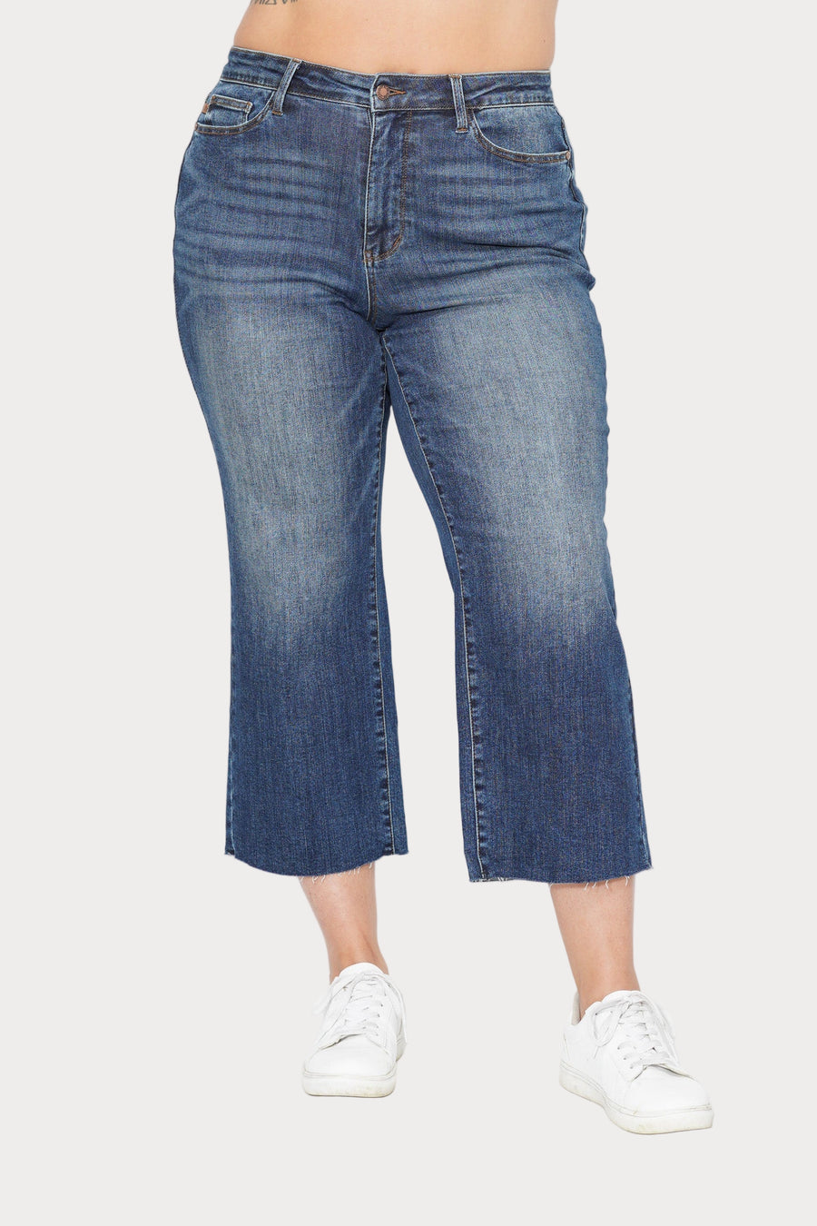 Front view of a plus-size model wearing the Dolly high-rise cropped wide-leg jeans in a medium wash with a raw hem. She is standing against a light grey background and wearing white sneakers.