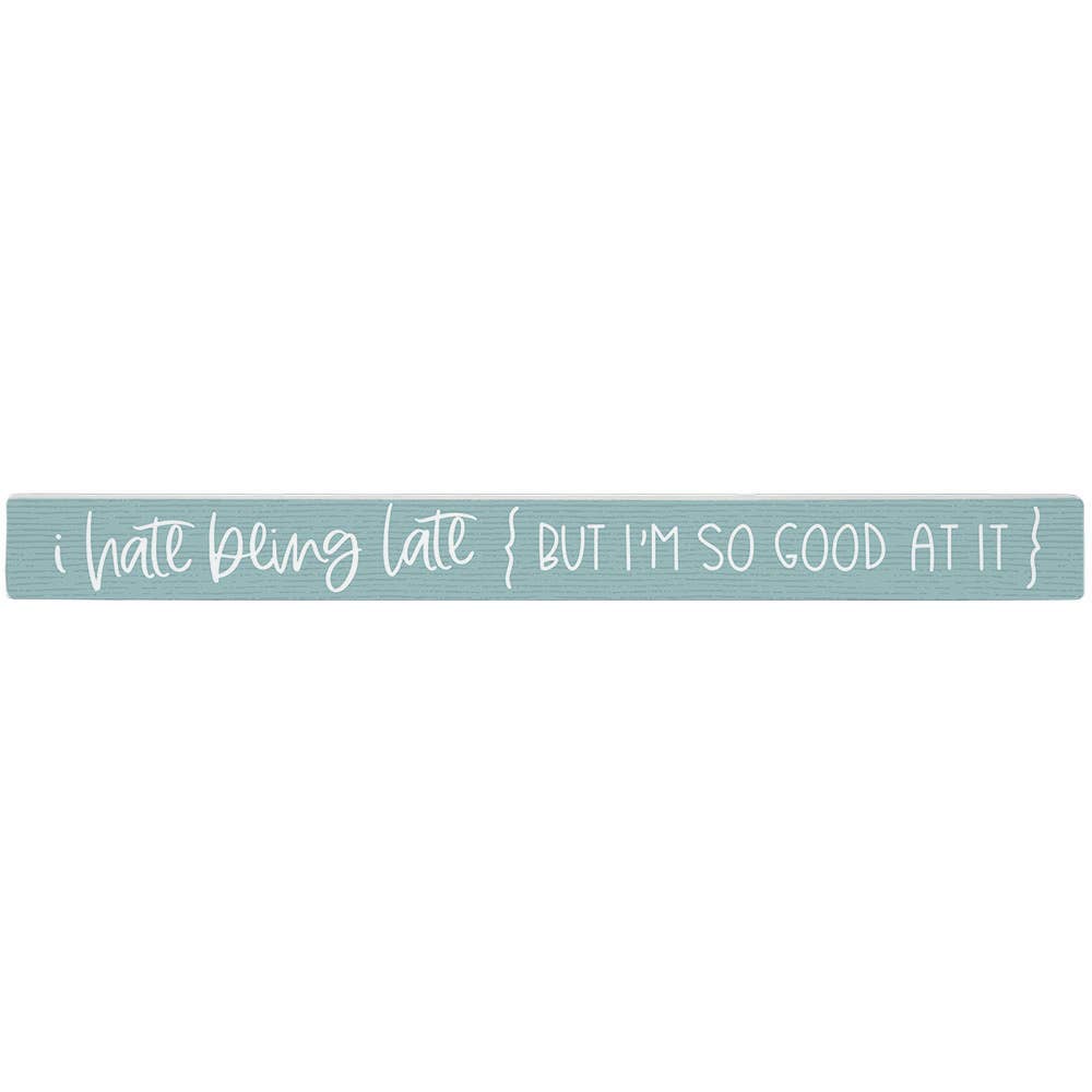 Textual phrase 'i hate being late { BUT I'M SO GOOD AT IT }' on a light blue wooden block. Shown on a white background
