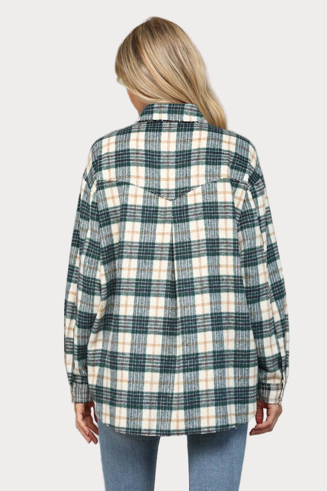 A woman wearing an oversized hunter green, cream, and tan plaid shacket with raw edge chest pockets and snap buttons, paired with light wash jeans.