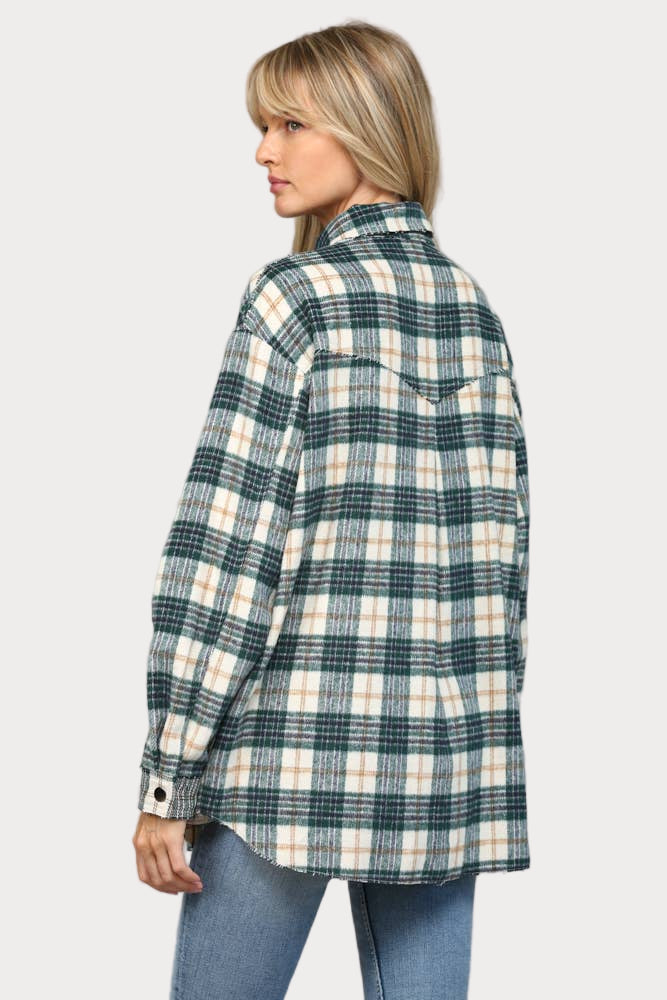 A woman wearing an oversized hunter green, cream, and tan plaid shacket with raw edge chest pockets and snap buttons, paired with light wash jeans.
