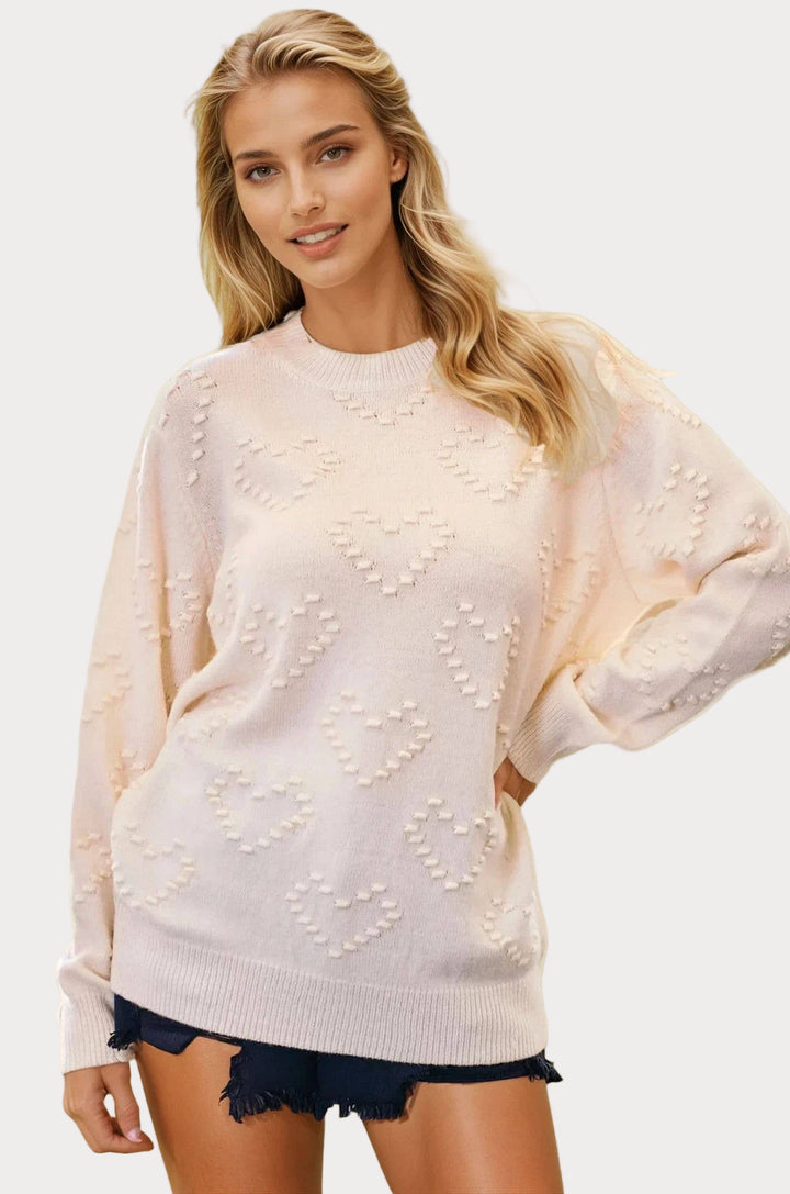 A female model wearing a cream knit sweater with a heart detail and long balloon sleeves, paired with denim shorts.