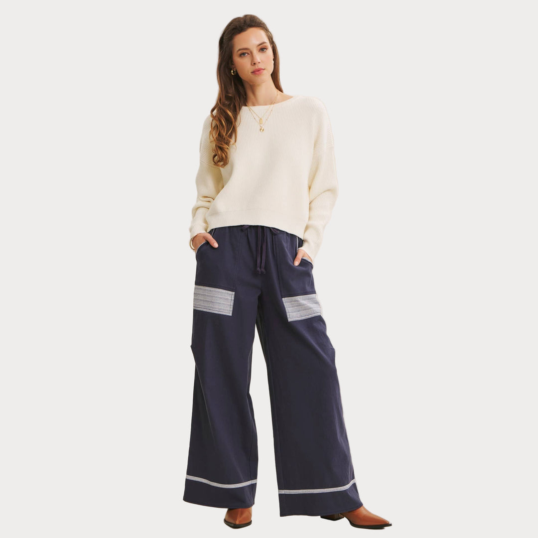 Model wearing Bow Back Sweater | Dolman Sleeve Knit with Satin Tie paired with navy pants with white lace patches and striped cuff on beige background