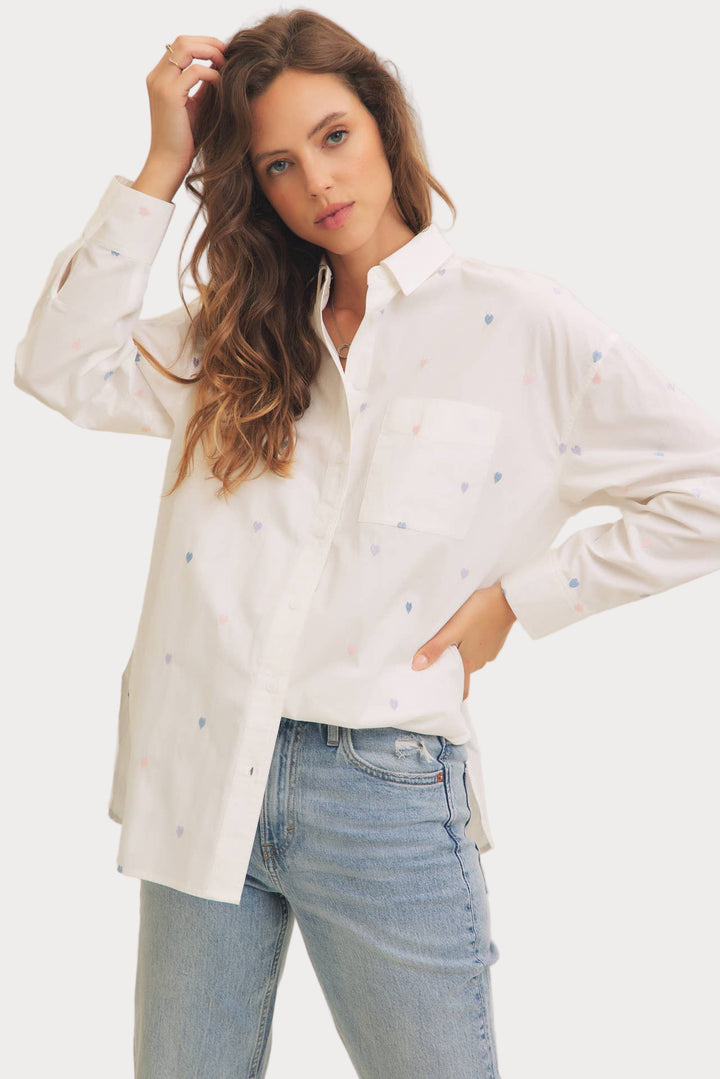 A woman wearing a white long-sleeved button-down shirt with heart embroidery and a collar, paired with blue jeans.