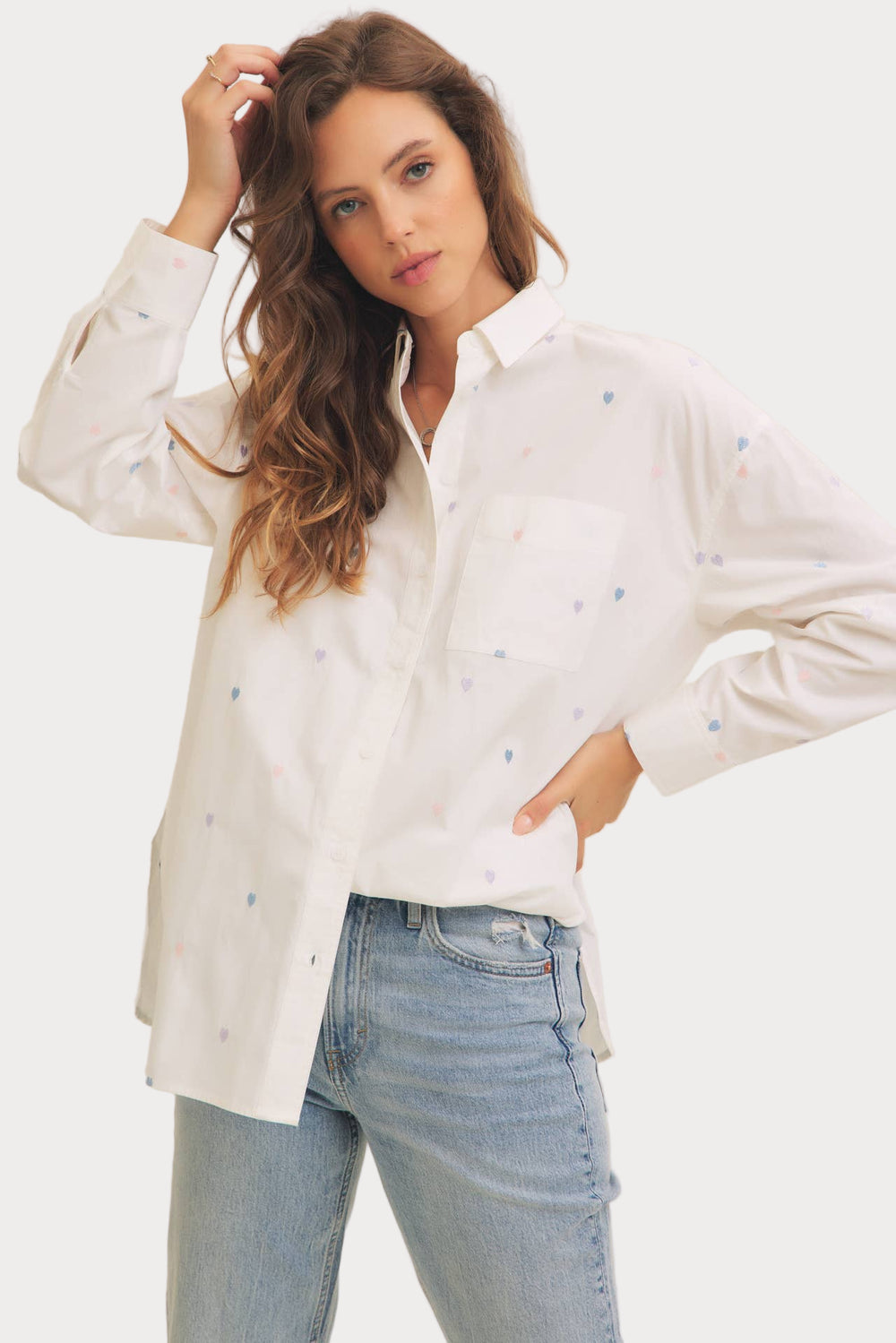 A female model wearing a long-sleeved, button-down shirt with heart embroidery in a pastel color, paired with blue jeans.