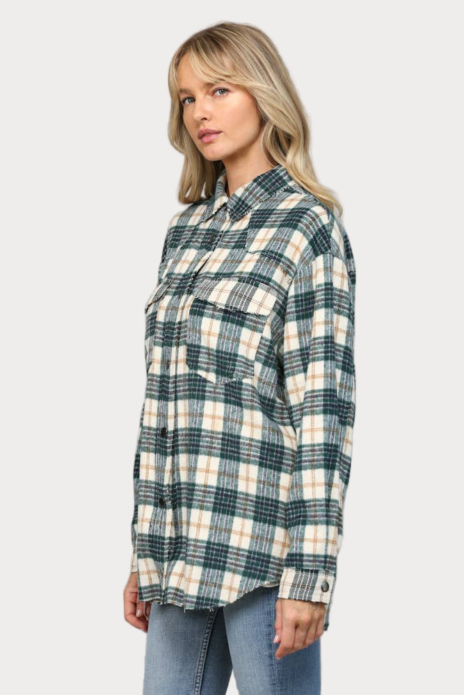 A woman wearing an oversized hunter green, cream, and tan plaid shacket with raw edge chest pockets and snap buttons, paired with light wash jeans.