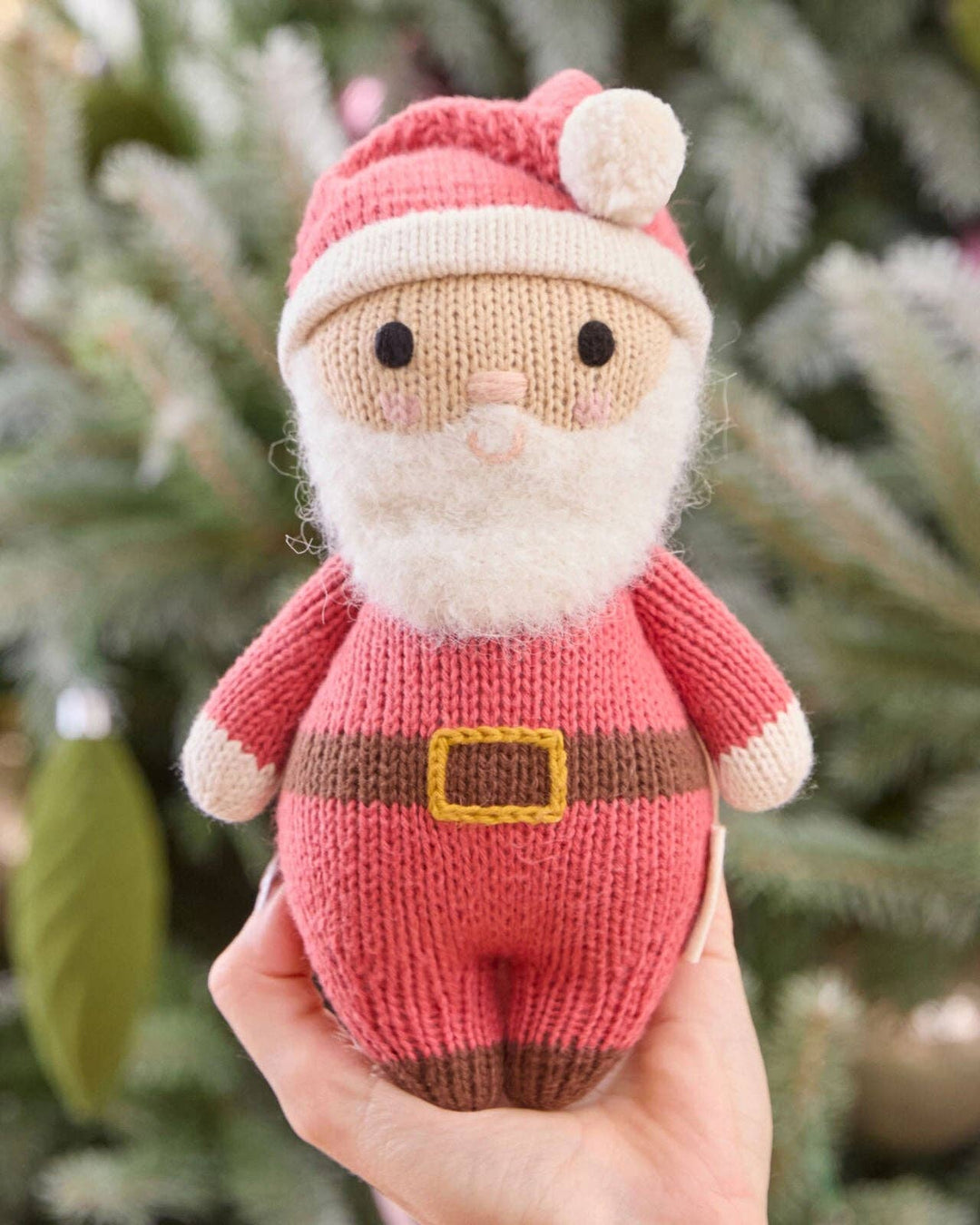 A hand holding a small knitted Santa Claus doll with a pink outfit, cream beard and hat, and brown belt, set against a blurred Christmas tree background.