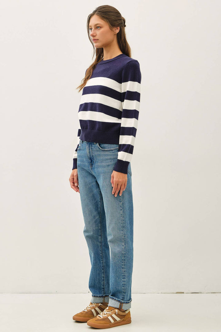A woman modeling a navy and white horizontally striped long-sleeve knit sweater, paired with light-wash blue jeans and brown sneakers, against a plain off-white studio background.