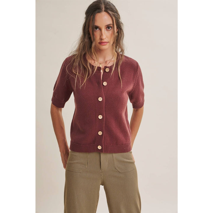 A woman modeling a short-sleeve button-front cardigan in burgundy over a beige tank top and brown stripped trousers, photographed against a plain light beige studio background.