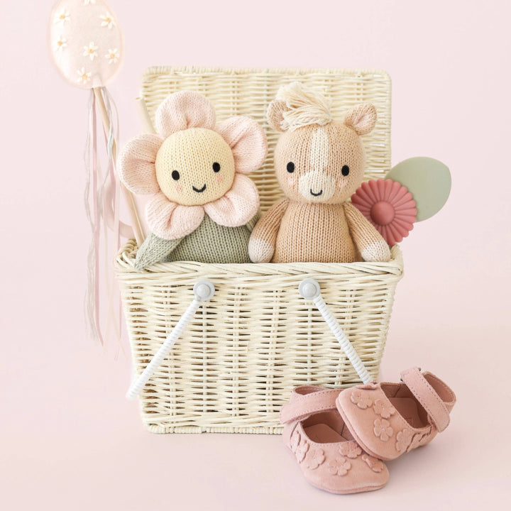 Wicker basket with knitted toys, including a flower and a horse, on a pink background.