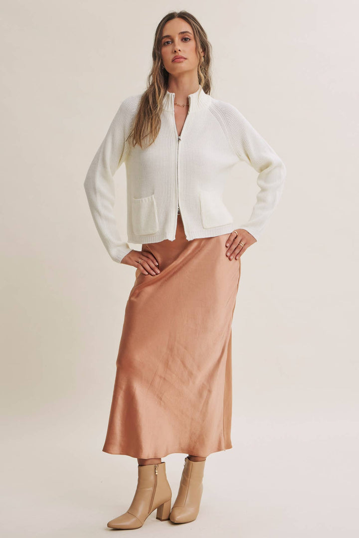 A woman modeling a white cropped zip-front jacket with front patch pockets, paired with a satin-like blush pink midi skirt and beige ankle boots, against a plain light beige studio background.