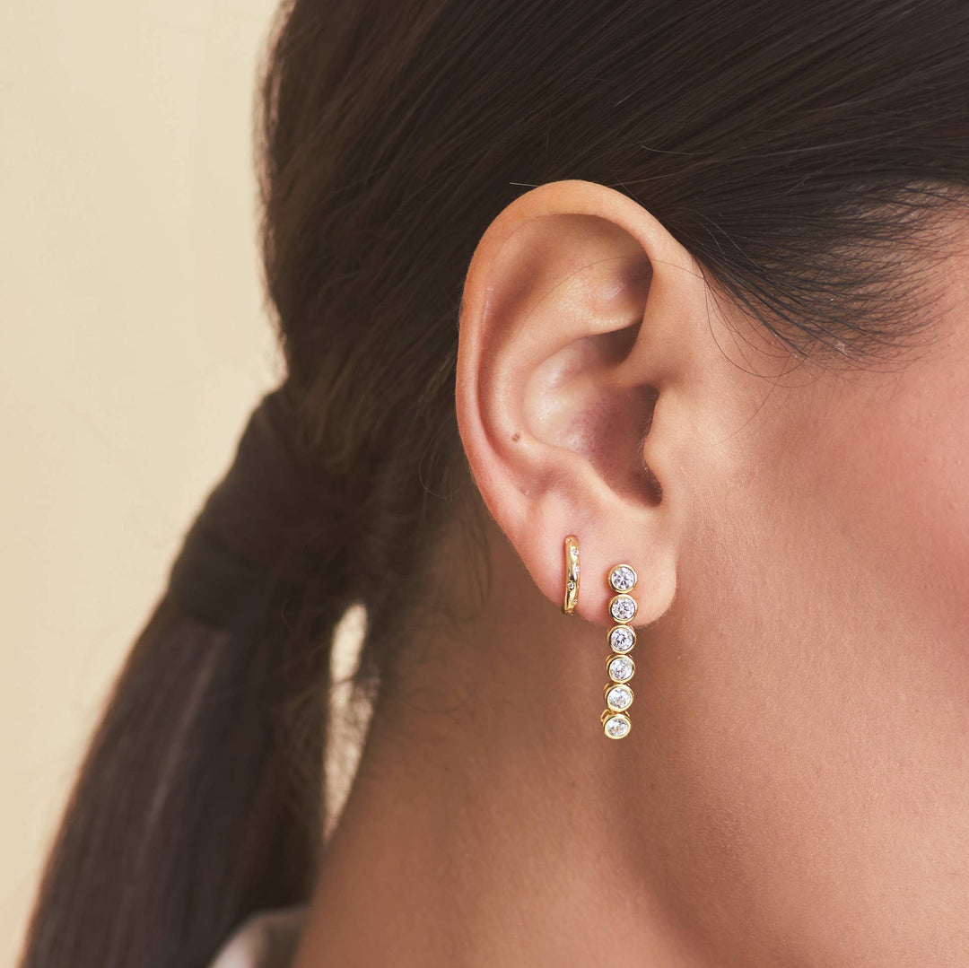 Close-up of an ear wearing a gold earring with clear gemstones against a neutral background