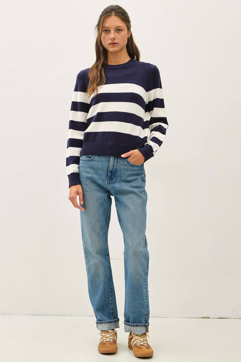 A woman modeling a navy and white horizontally striped crewneck sweater, paired with light-wash blue jeans and brown suede sneakers, against a plain white studio background.