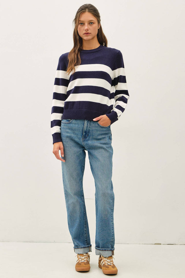 A woman modeling a navy and white horizontally striped crewneck sweater, paired with light-wash blue jeans and brown suede sneakers, against a plain white studio background.