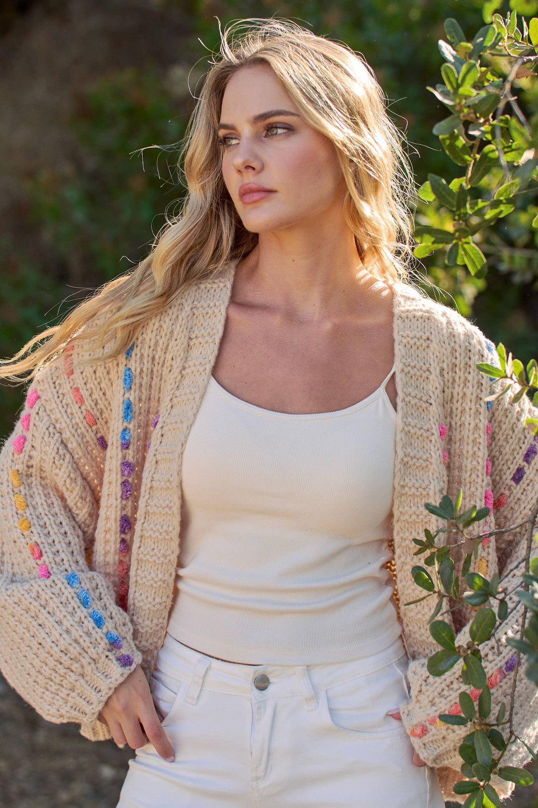 A woman outdoors wearing a cream-colored chunky knit cardigan with colorful embroidered details on the sleeves, layered over a white tank top and white pants.