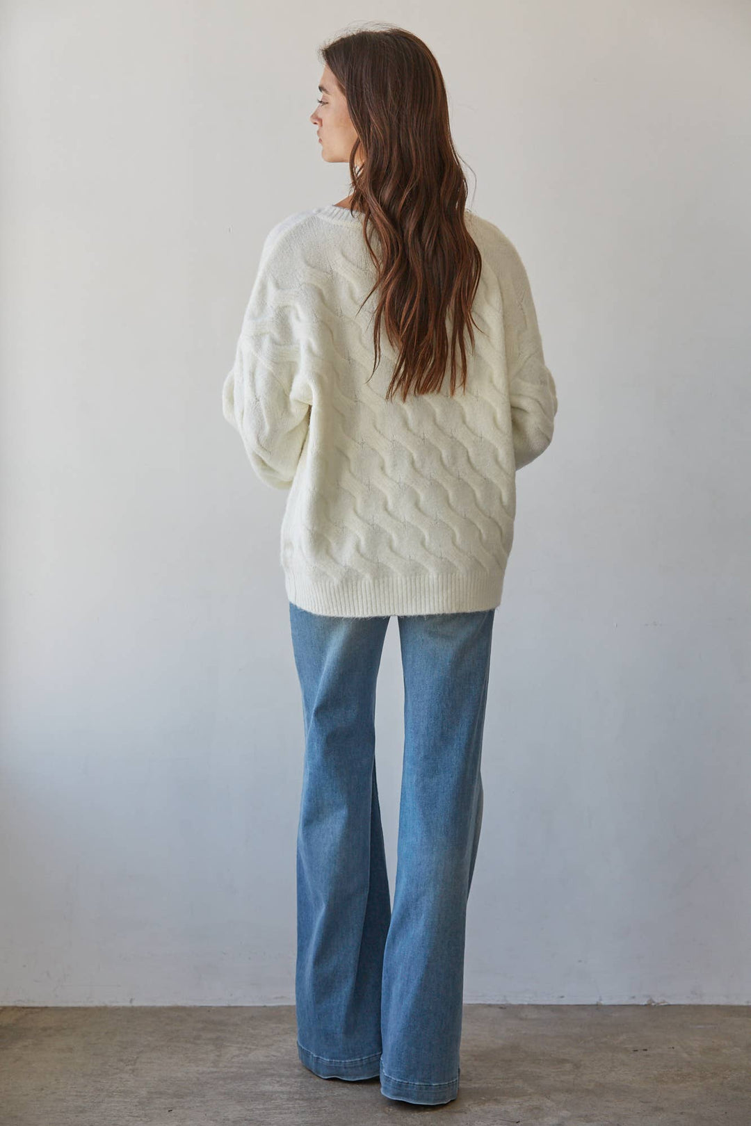 A woman is shown from behind wearing a cream-colored cable-knit sweater and blue jeans, standing against a plain light gray wall.