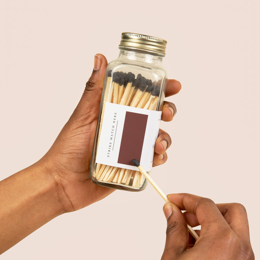 Hand holding a glass matchbox with matches and a lit match, against a beige background