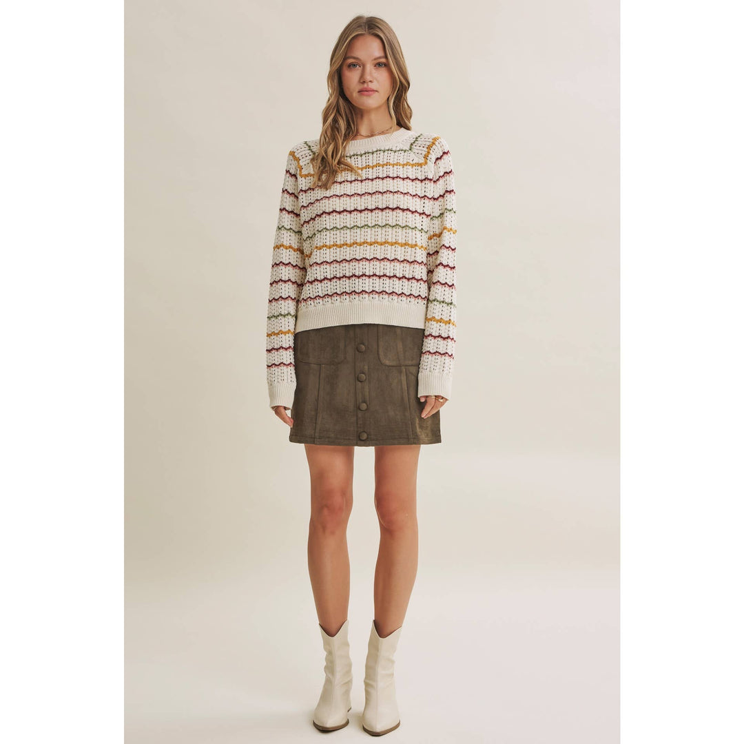 A woman modeling a cream knit sweater with horizontal multicolor stripes in red, yellow, and brown, paired with a dark brown mini skirt and white ankle boots, against a solid light beige background.