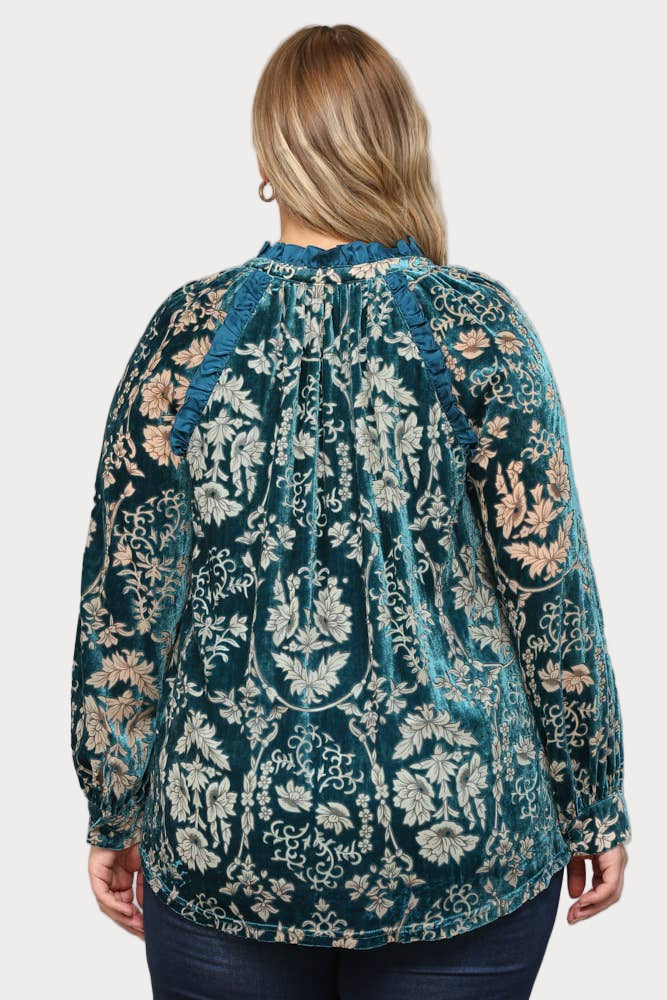 A smiling plus-size model wears a long-sleeve teal burnout velvet blouse featuring a floral pattern, ruffle trim, and a tassel tie at the neck.

