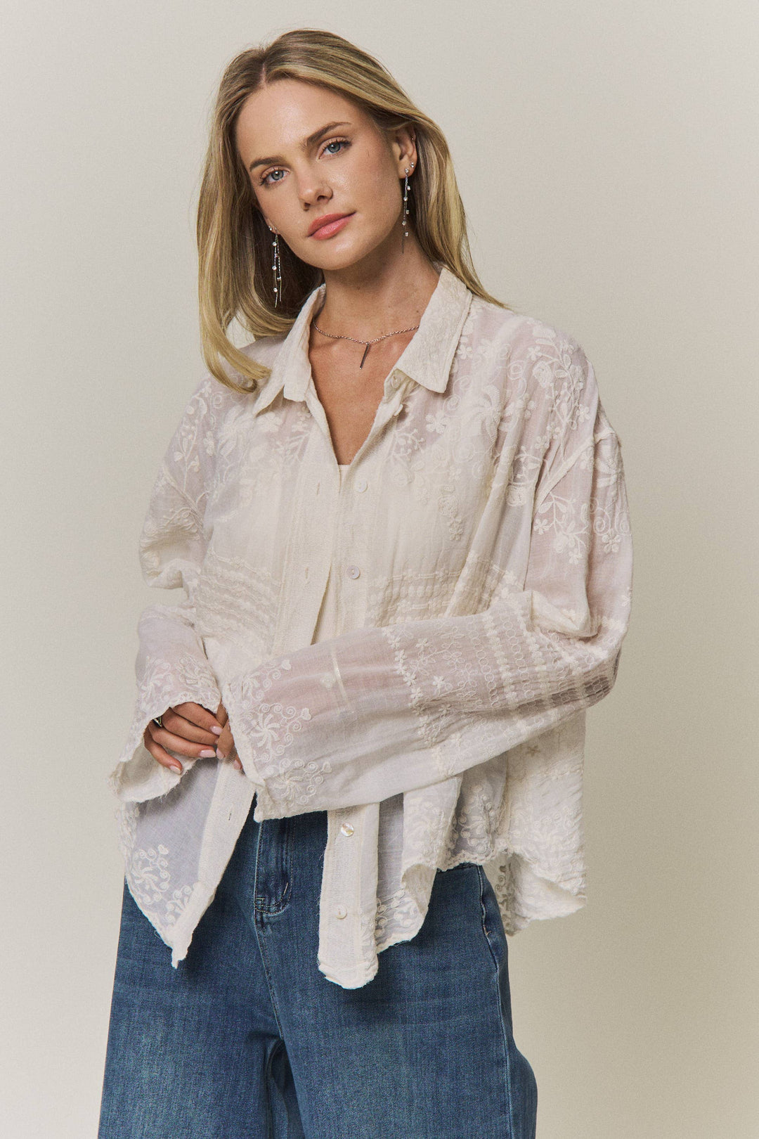 Woman wearing a light-colored lace embroidered blouse and blue jeans against a plain background