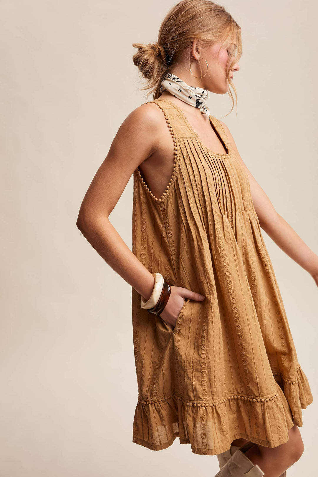 Woman wearing a textured camel-colored sleeveless mini dress with pintuck details and ruffle hem. Shows pocket detail