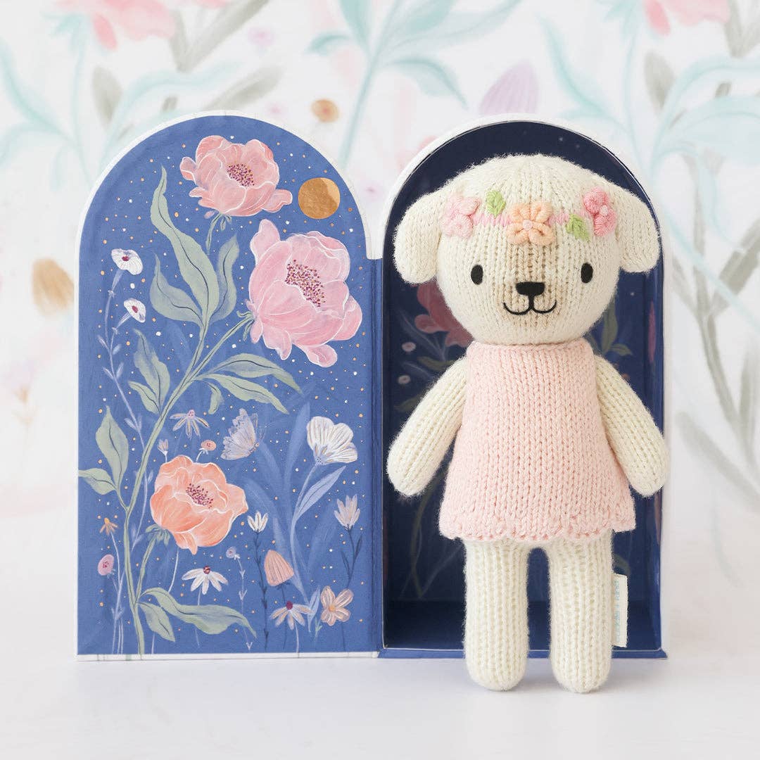 Knitted dog in a pink dress with floral decorations, standing in front of a floral-themed box.
