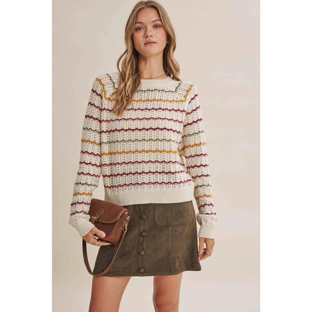 A woman modeling a cream knit sweater with horizontal multicolor stripes in yellow, red, and green, paired with an olive green mini skirt and a brown leather crossbody bag, set against a solid beige studio background.