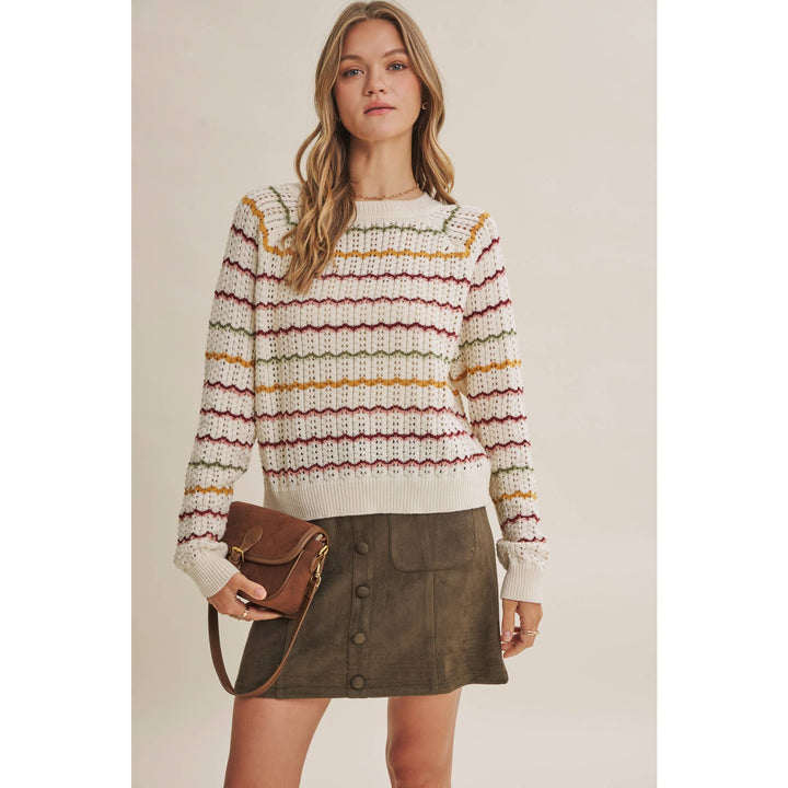 A woman modeling a cream knit sweater with horizontal multicolor stripes in yellow, red, and green, paired with an olive green mini skirt and a brown leather crossbody bag, set against a solid beige studio background.