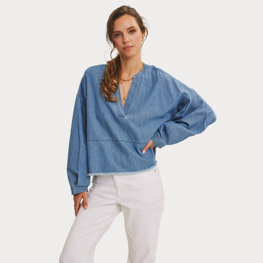 Woman wearing a relaxed fit denim V-neck top with frayed hem, styled with white pants for a casual chic look.