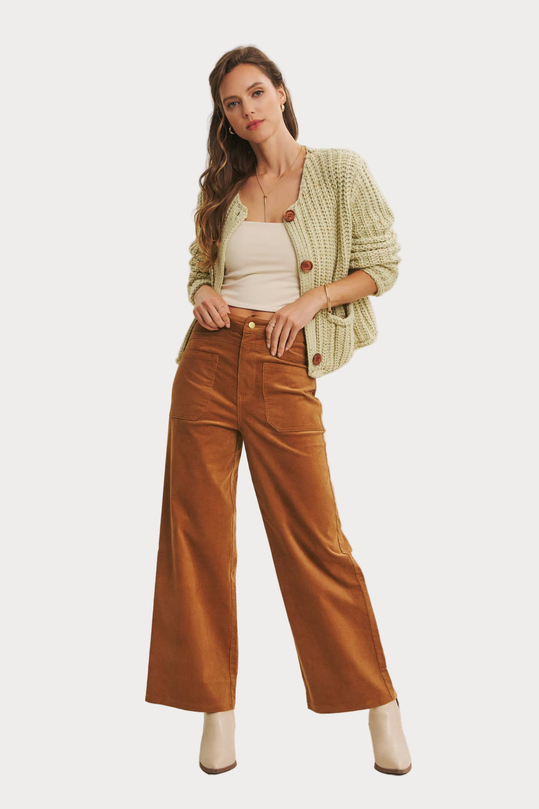 Woman wearing a green cardigan, white top, brown wide-leg pants, and beige boots on a plain background