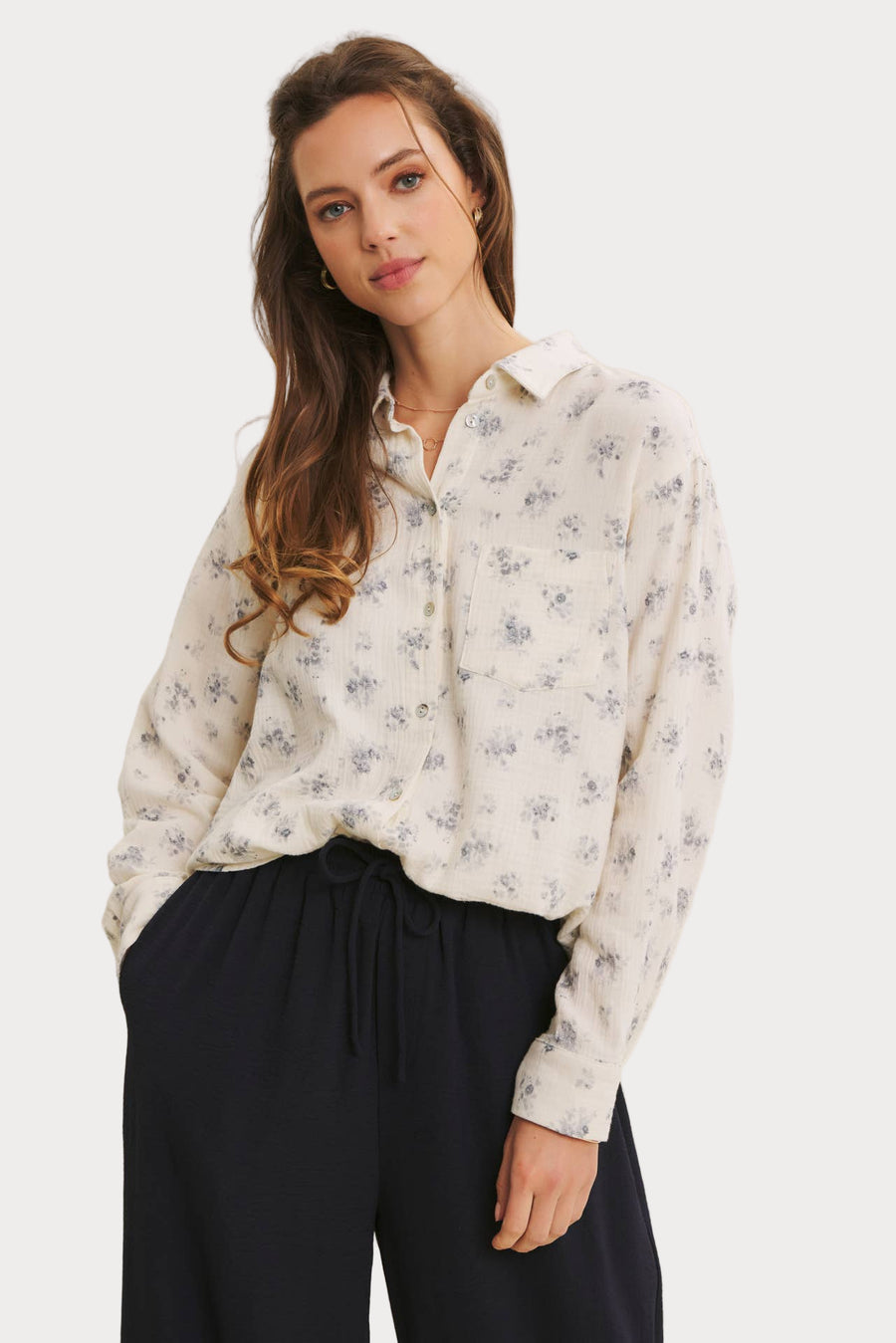 Woman wearing a white floral blouse and dark pants against a plain background