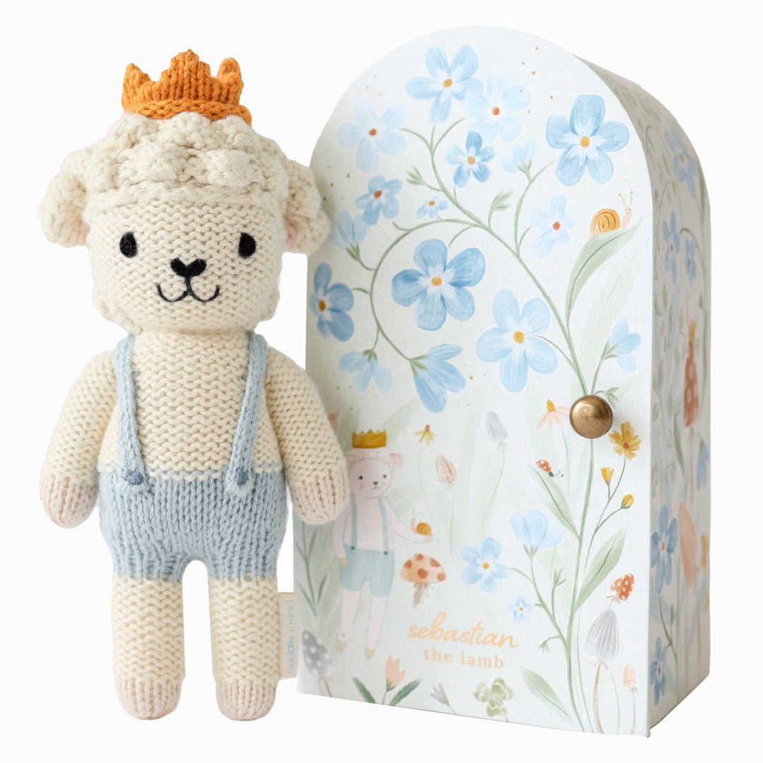 Knitted toy lamb with a crown and overalls next to a floral-patterned box