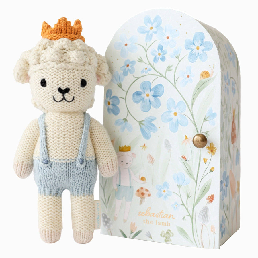 Knitted toy lamb with a crown and overalls next to a floral-patterned box