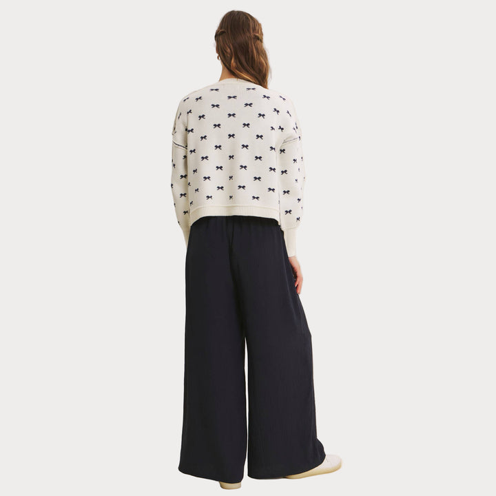 Model wearing an ivory jacquard knit sweater with a repeating navy blue bow pattern and voluminous balloon sleeves. Paired with wide-leg navy pants and white sneakers, styled for a casual, chic look. Model showing back profile.

