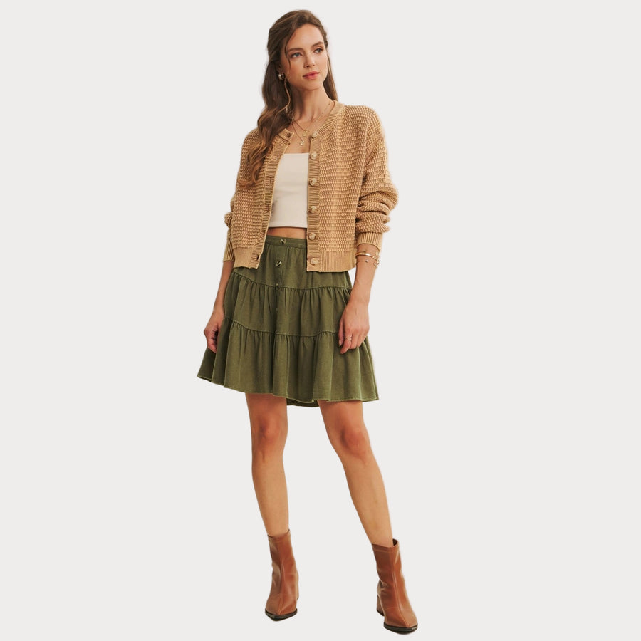 Woman wearing a beige cardigan, white top, green skirt, and brown boots on a plain background