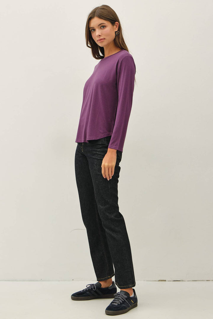 A woman modeling a purple long-sleeve crewneck top, paired with black jeans and dark blue sneakers, standing against a plain off-white studio background.