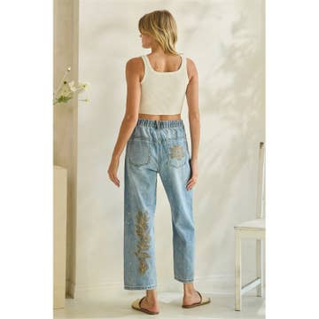 Woman wearing a white tank top and light blue jeans with floral embroidery in a room. back view.