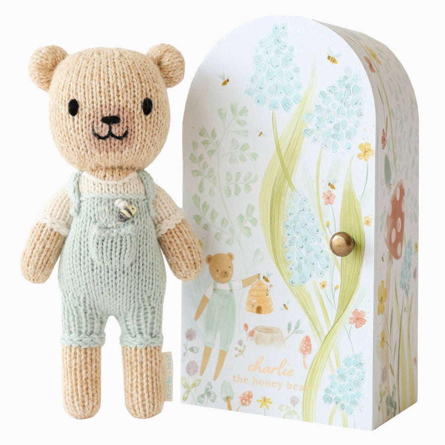 Knitted teddy bear in overalls next to its packaging with floral design