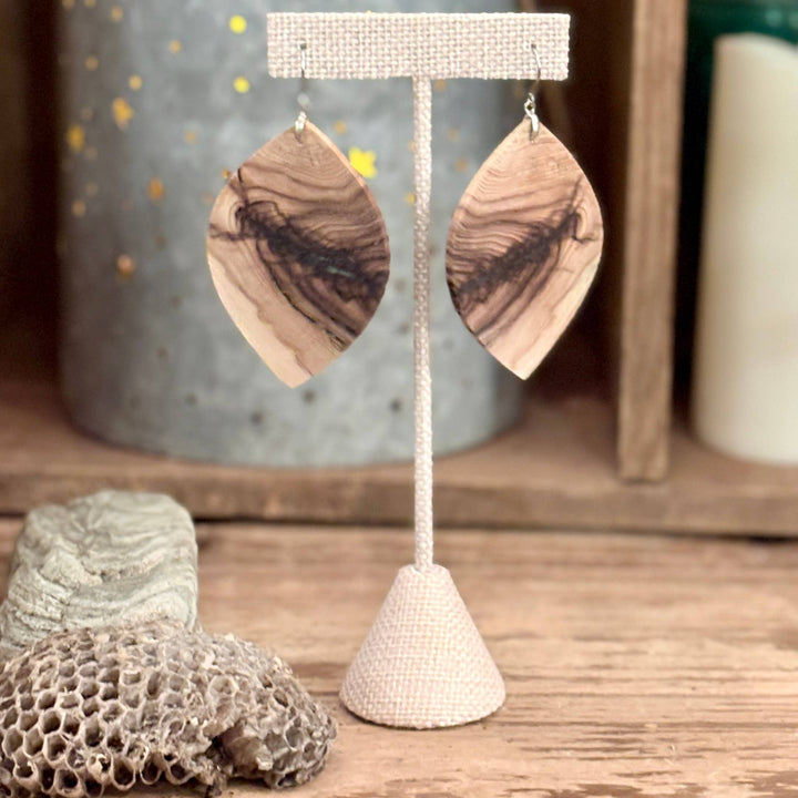 Wooden leaf-shaped earrings on a stand with a textured candle and decorative items in the background.