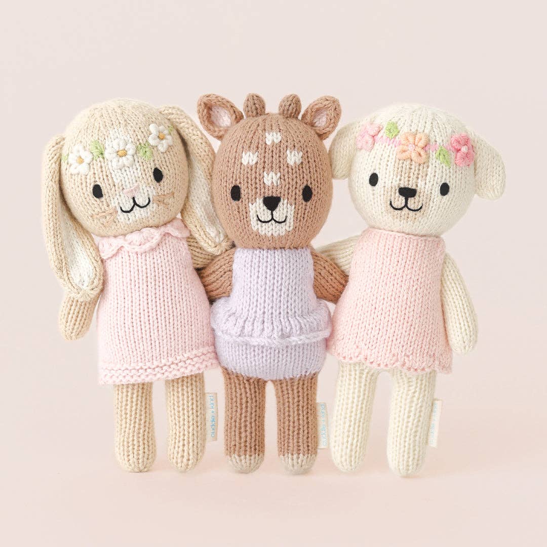 Three knitted toys resembling a rabbit, deer, and dog with floral hair accessories on a light pink background.