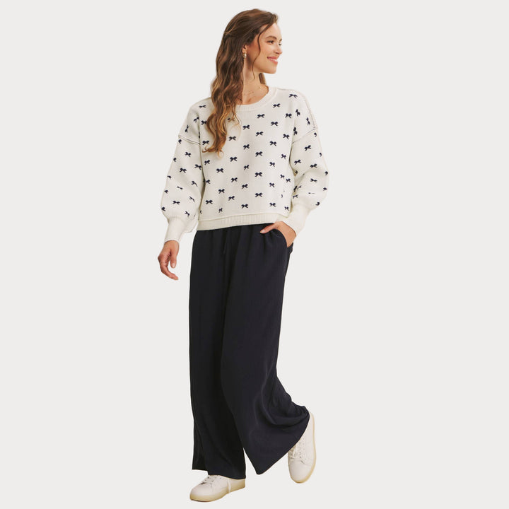 Model wearing an ivory jacquard knit sweater with a repeating navy blue bow pattern and voluminous balloon sleeves. Paired with wide-leg navy pants and white sneakers, styled for a casual, chic look. 

