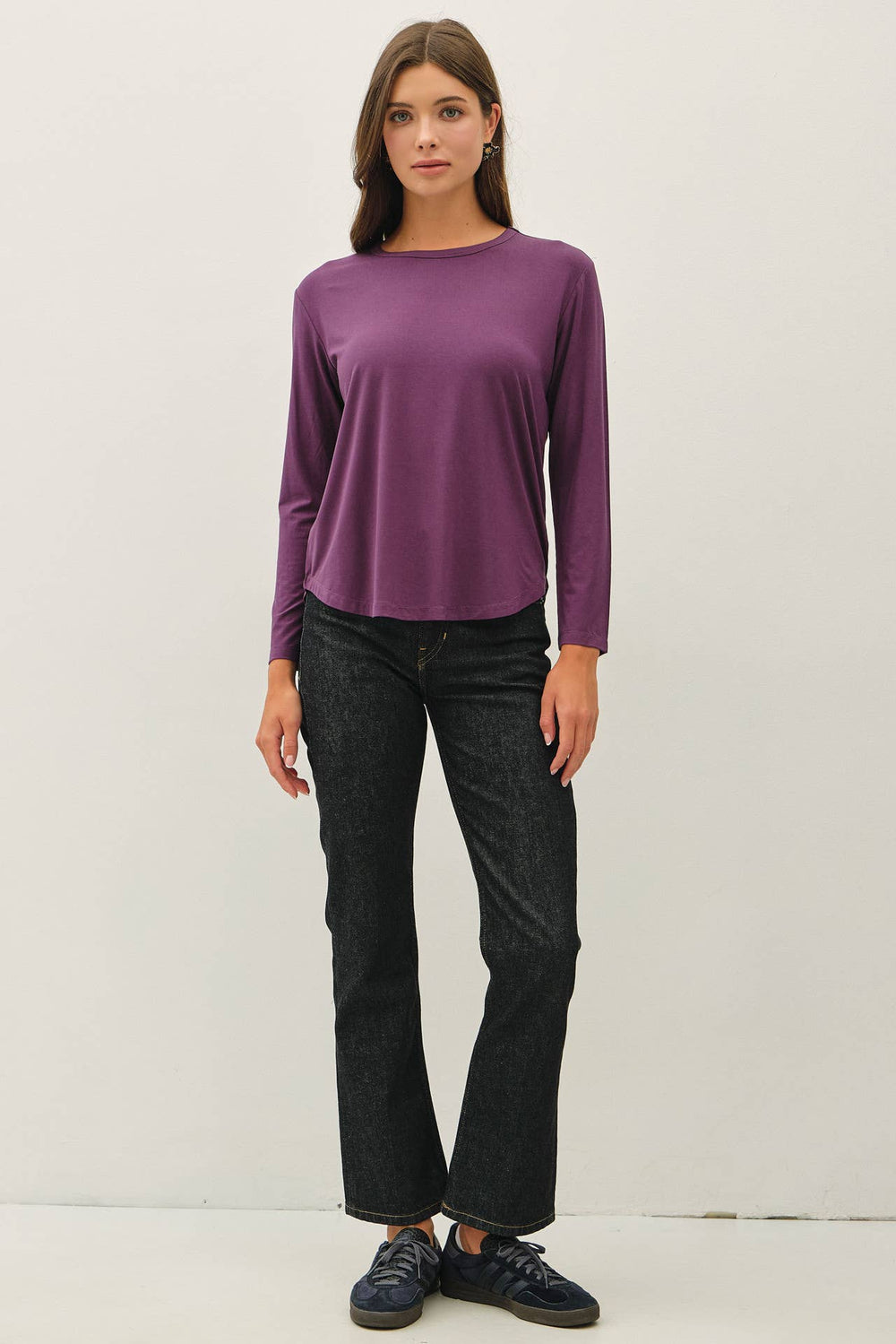 A woman modeling a long-sleeve crewneck top in a muted purple color, paired with dark wash jeans and dark sneakers, standing against a plain off-white studio background.