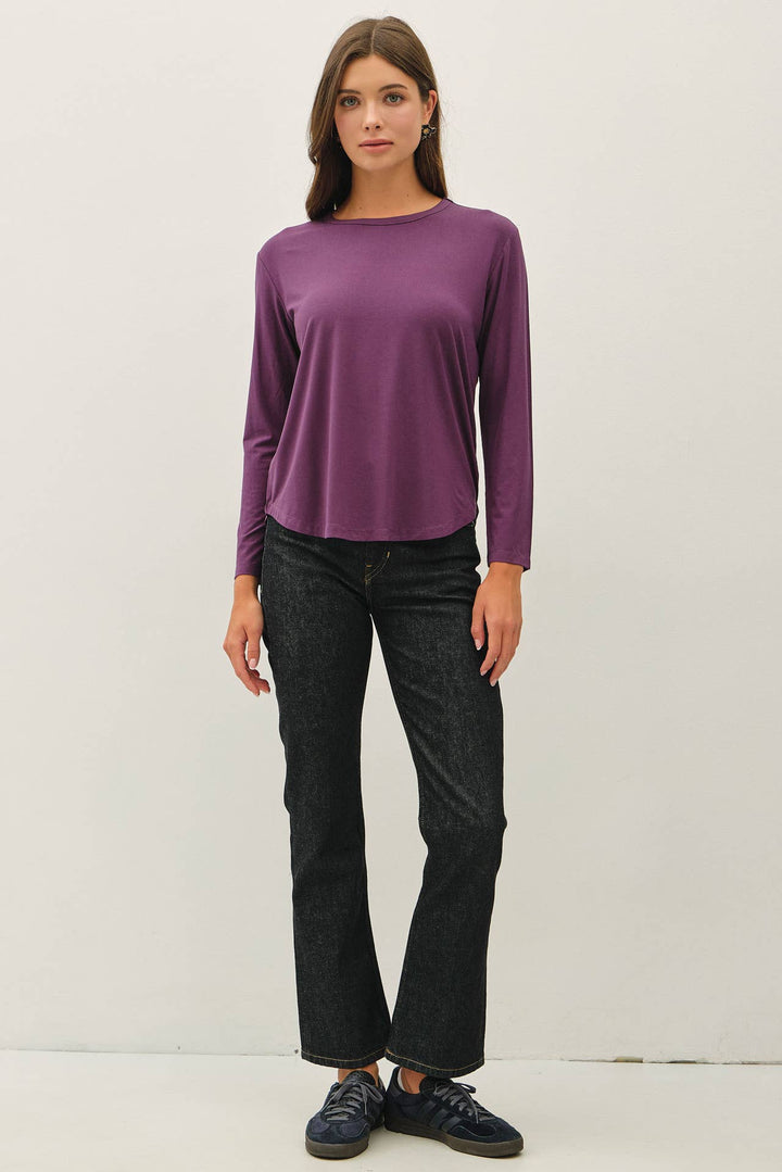 A woman modeling a long-sleeve crewneck top in a muted purple color, paired with dark wash jeans and dark sneakers, standing against a plain off-white studio background.