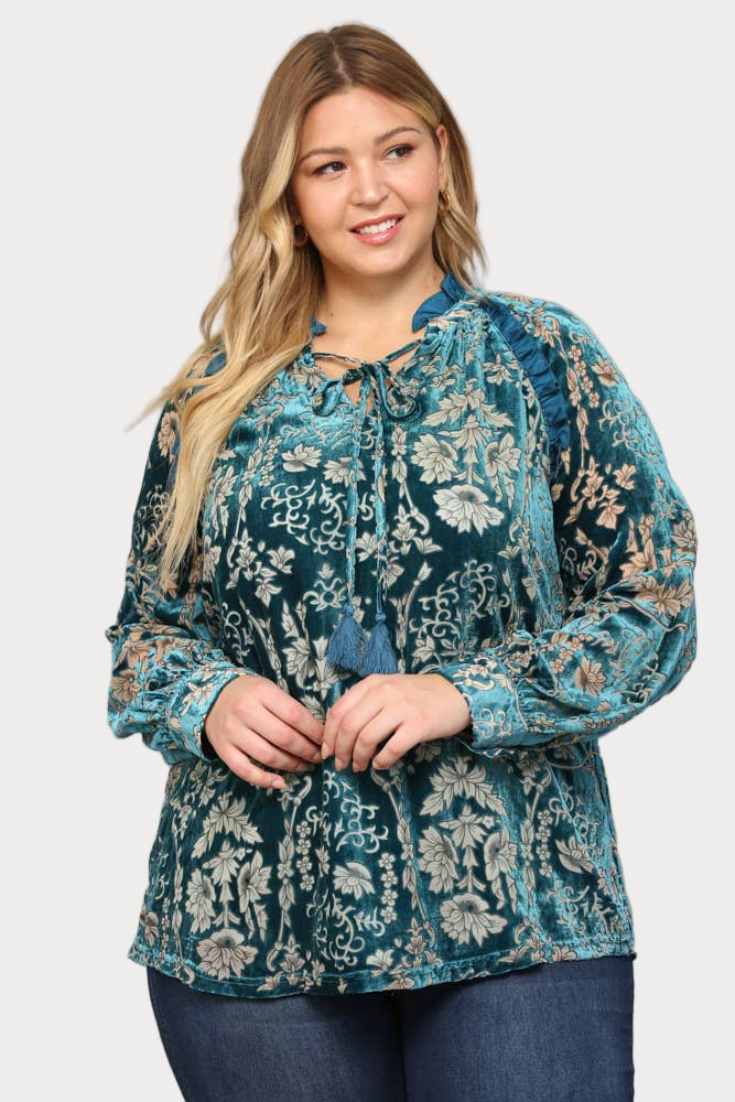 A smiling plus-size model wears a long-sleeve teal burnout velvet blouse featuring a floral pattern, ruffle trim, and a tassel tie at the neck.

