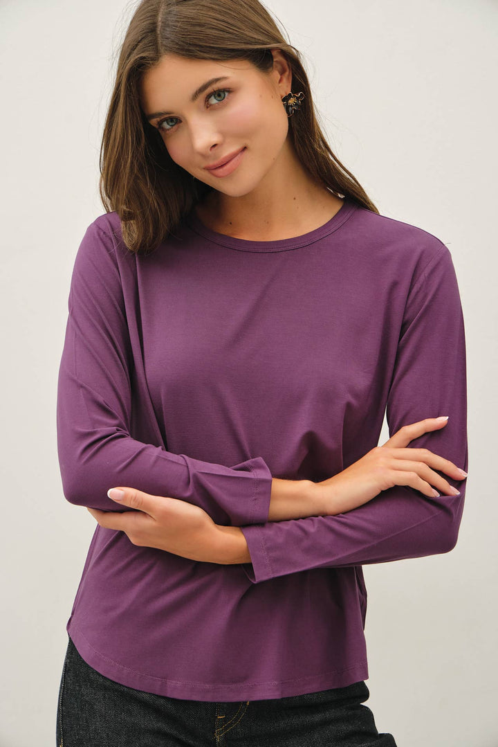 A woman wearing a solid purple long-sleeve crewneck top, posing with her arms crossed. The background is a clean, neutral off-white studio wall.