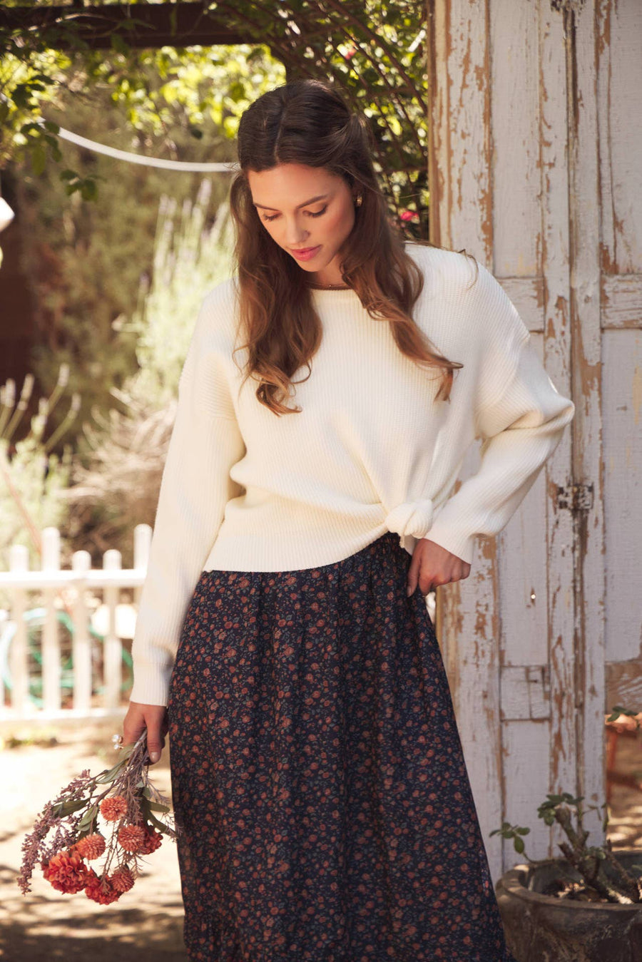 Model wearing Bow Back Sweater | Dolman Sleeve Knit with Satin Tie. Lifestyle photo with outdoor garden setting. Model posed looking down.
