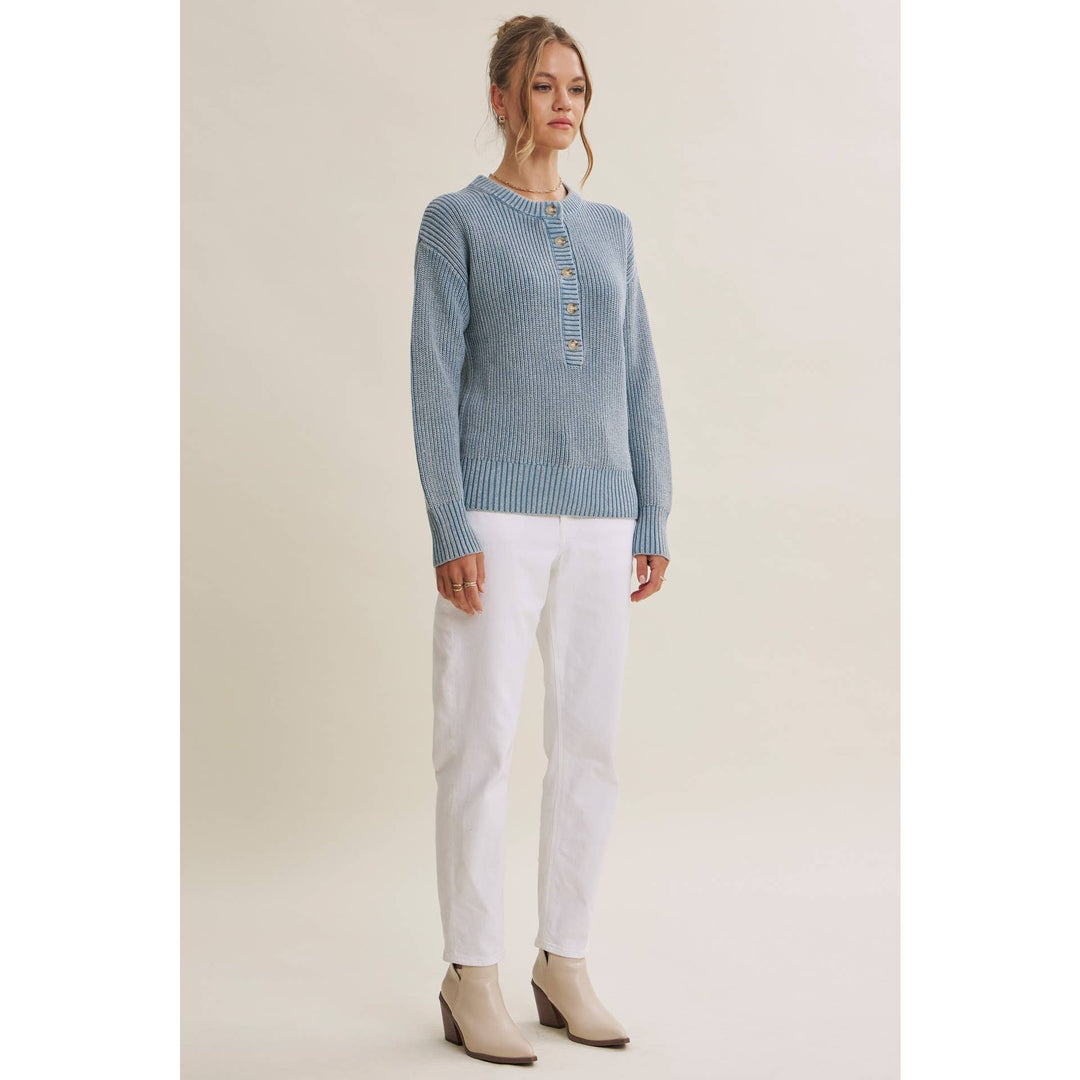 A woman modeling a light blue ribbed knit sweater with a buttoned henley neckline, paired with white pants and beige ankle boots, against a plain beige studio background.