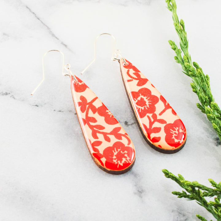 Teardrop-shaped earrings with red floral pattern on a marble surface with greenery.