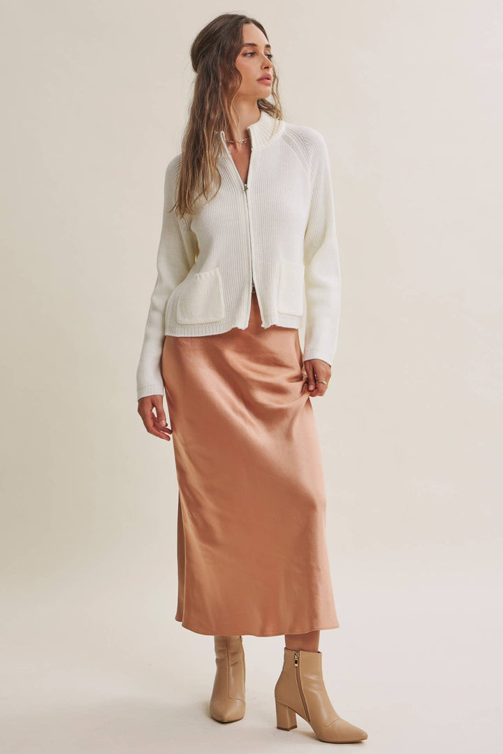 A woman modeling a white zip-up cardigan and a peach satin midi skirt with beige ankle boots, set against a plain beige studio background.